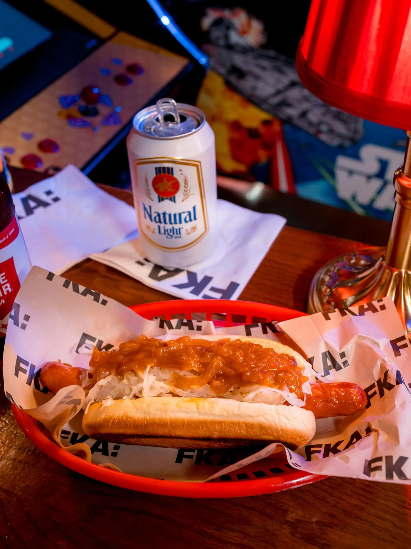 A hot dog with sauerkraut and onion sauce in a red basket, with arcade machines glowing behind it at FKA: Formerly Known As