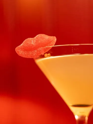 A pale martini in a conical glass with a sugared candy-lips garnish against a red background at FKA: Formerly Known As